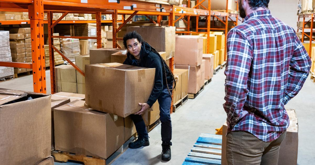 man lifting box in warehouse as supervisor watches